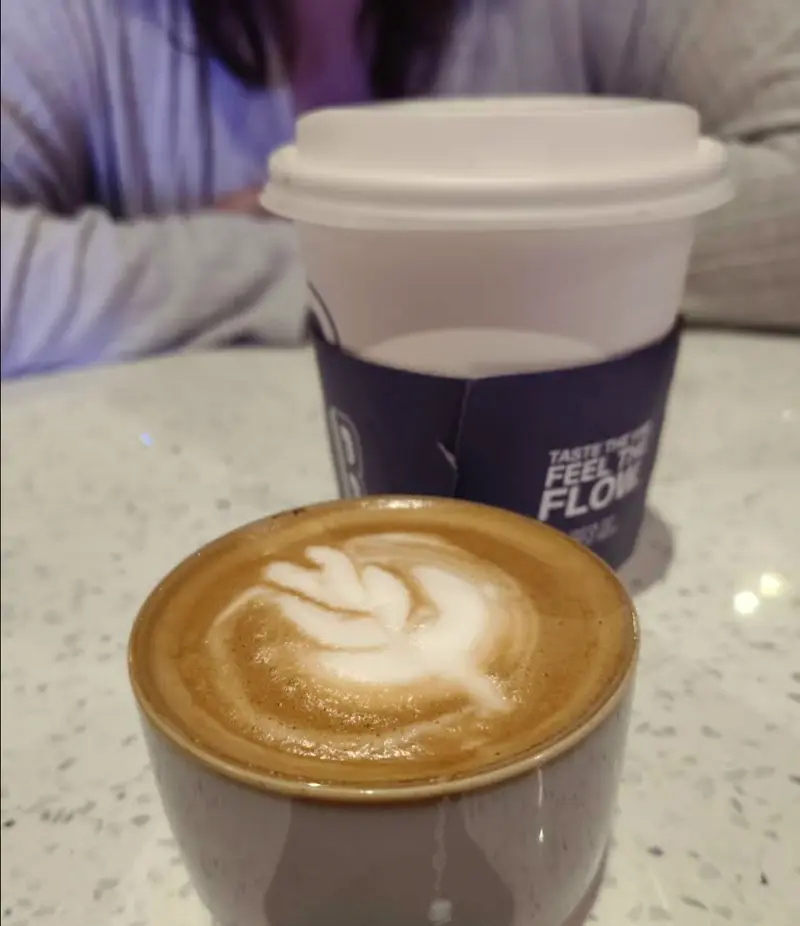 Latte con arte de hoja en taza gris junto a un vaso con la manga azul “Feel the flow”