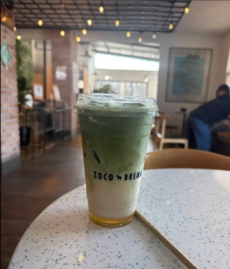Matcha latte frío en una mesa de terrazo con muro de ladrillo y focos colgantes al fondo