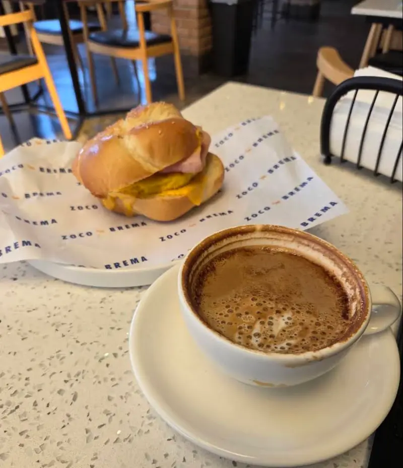 Mesa de terrazo con sándwich sobre el papel de Zoco Brema y capuchino; sillas de madera alrededor